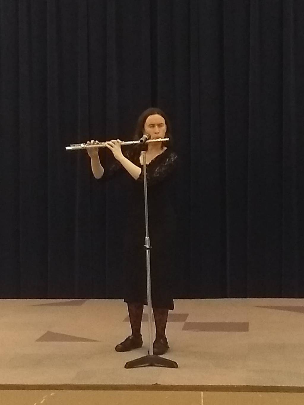 I, a European-presening white woman, am wearing a black dress playing flute. You can see the stage, a microphone I'm playing into, and a black curtain behind me.