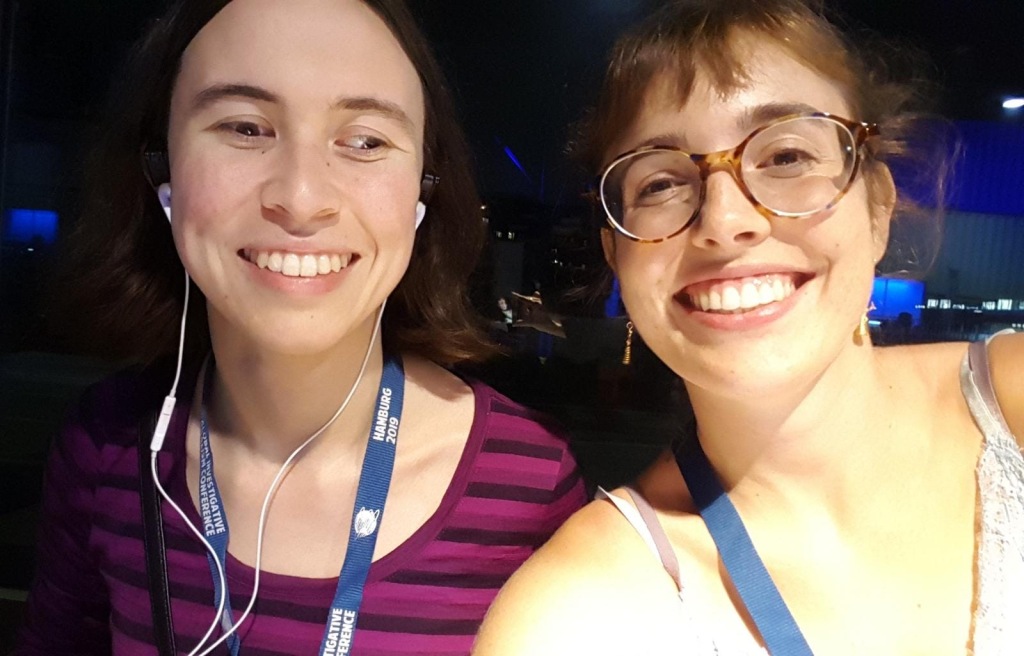 Two young European-presenting women, Ariadna and I, are sitting at a table smiling. We have blue Conference lanyards on, mine's facing text-side-out and reads "Hamburg 2019 / Global Investigative Journalism Conference".