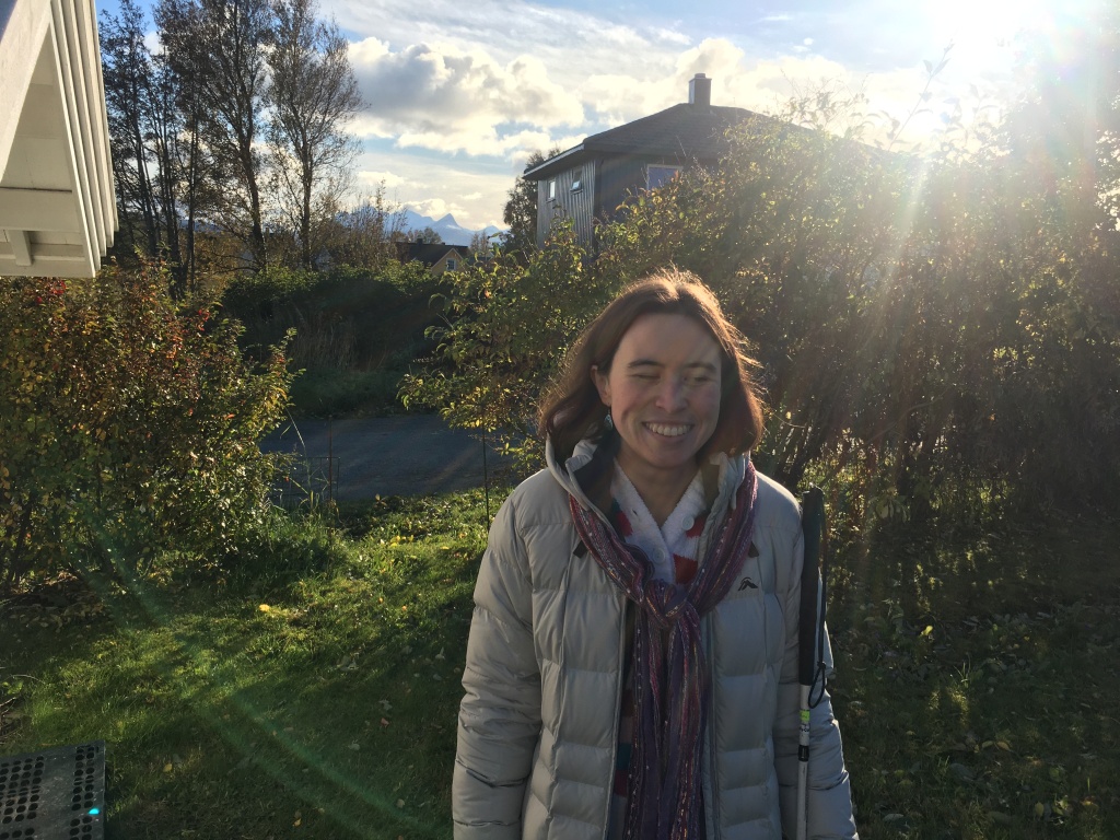 I'm standing on the grass behind my house on an autumn day, and you can see Norwegian mountains behind me. I'm a white woman with brown hair, wearing a puffy winter jacket and multi-colour scarf.